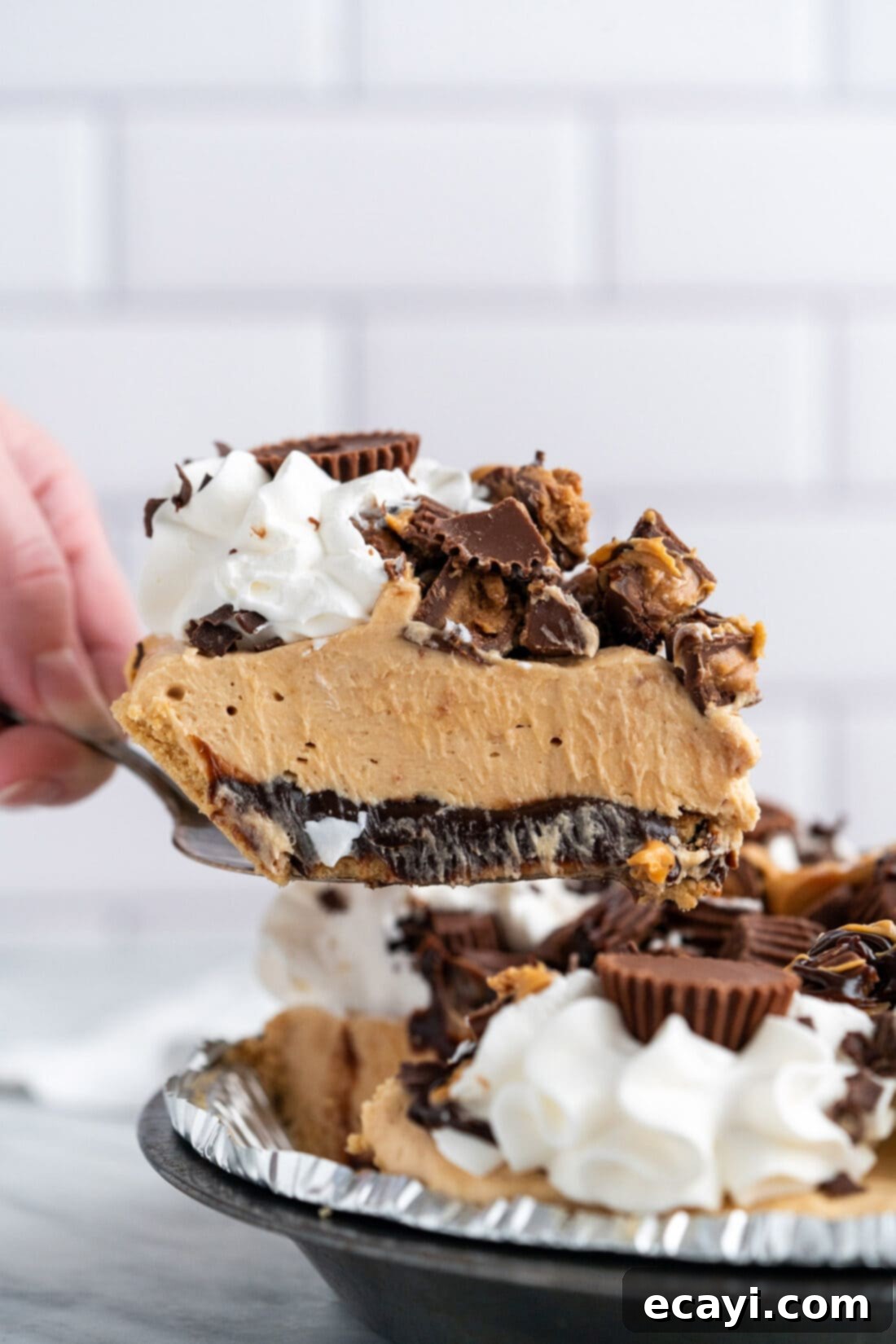 Peanut Butter Perfection Pie 2 A glorious slice of No-Bake Reese's Peanut Butter Pie being removed from the pan, revealing its distinct layers of graham cracker crust, fudge, and peanut butter filling, topped with whipped cream and chopped Reese's.