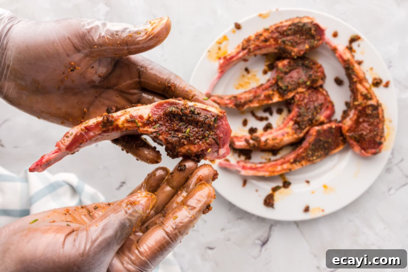 Tender Oven-Baked Lamb Chops 7 Hands wearing gloves massaging the spice rub onto lamb chops on a cutting board.