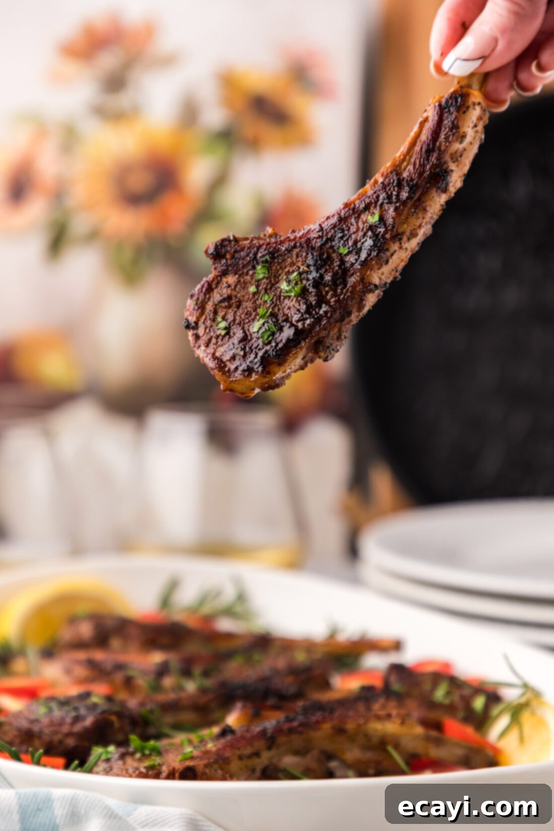 Tender Oven-Baked Lamb Chops 10 A hand holding a beautifully seared and baked lamb chop, showcasing its tender interior, with a blurred background of a full platter.