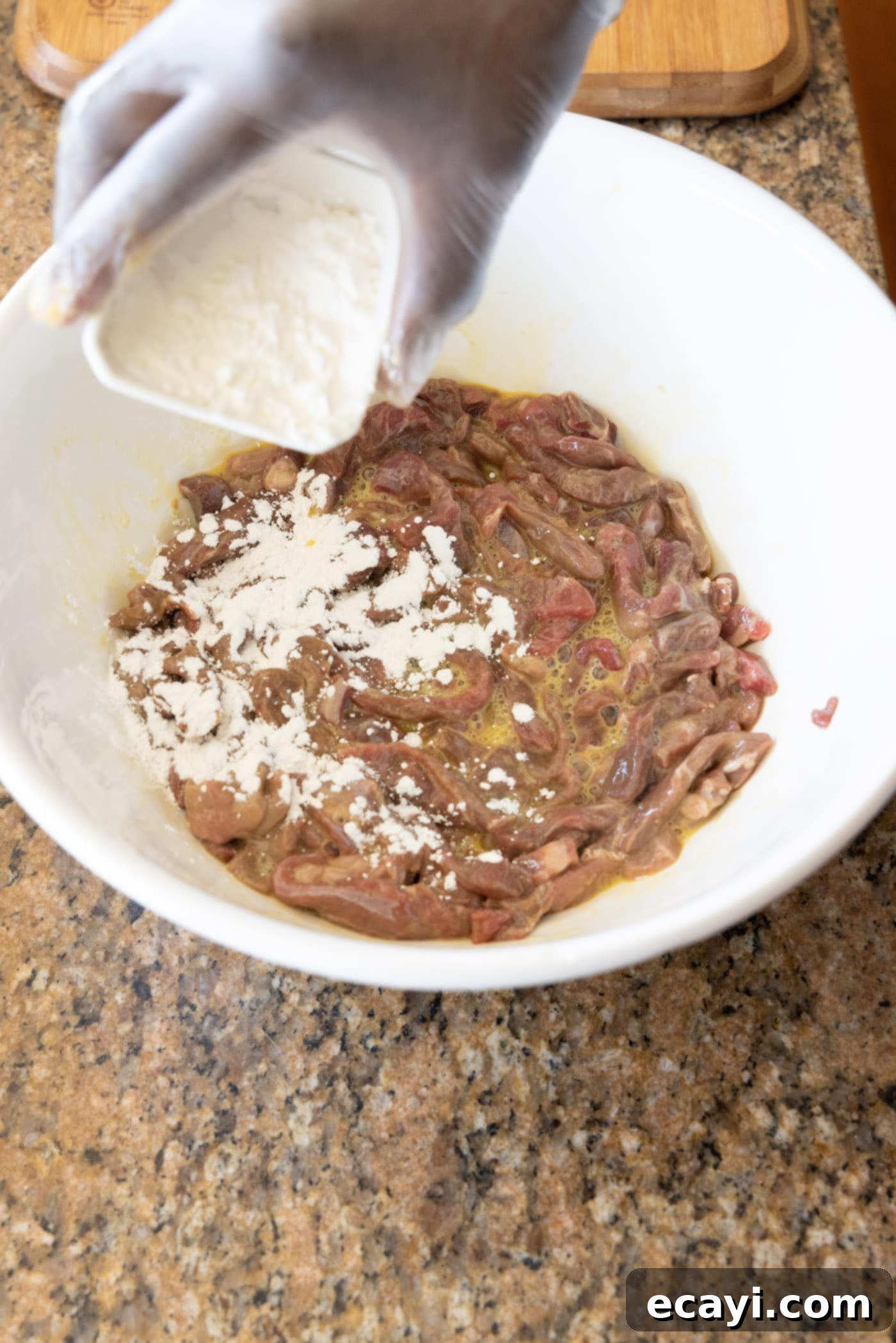 Crispy Beijing Beef 7 Gloved hands mixing cornstarch into the egg-coated beef strips in a large bowl, ensuring even coverage.