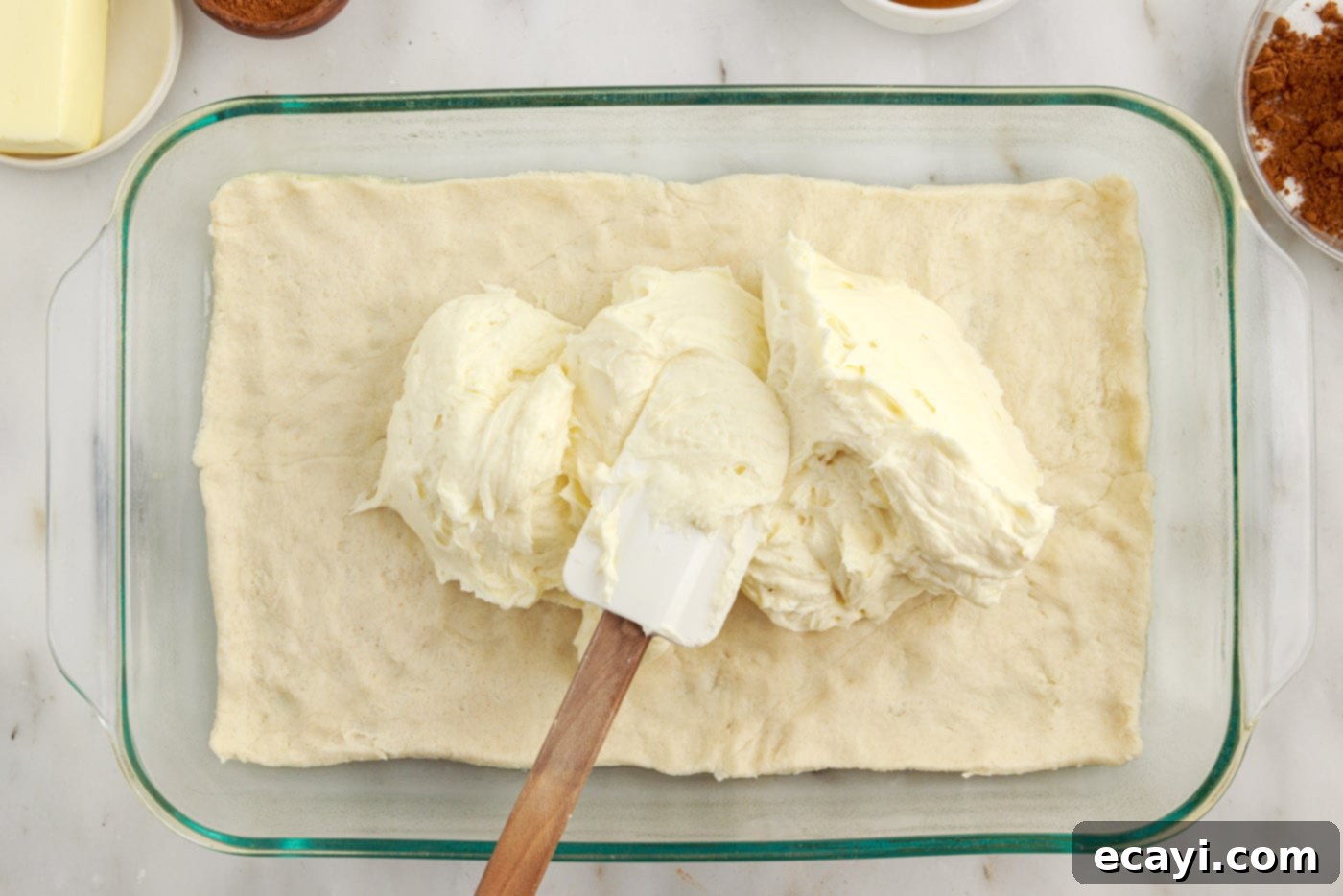 Cinnamon Swirl Sopapilla Cheesecake 8 A spatula gently spreading the creamy cheesecake filling in an even layer over the crescent dough base in a baking dish.