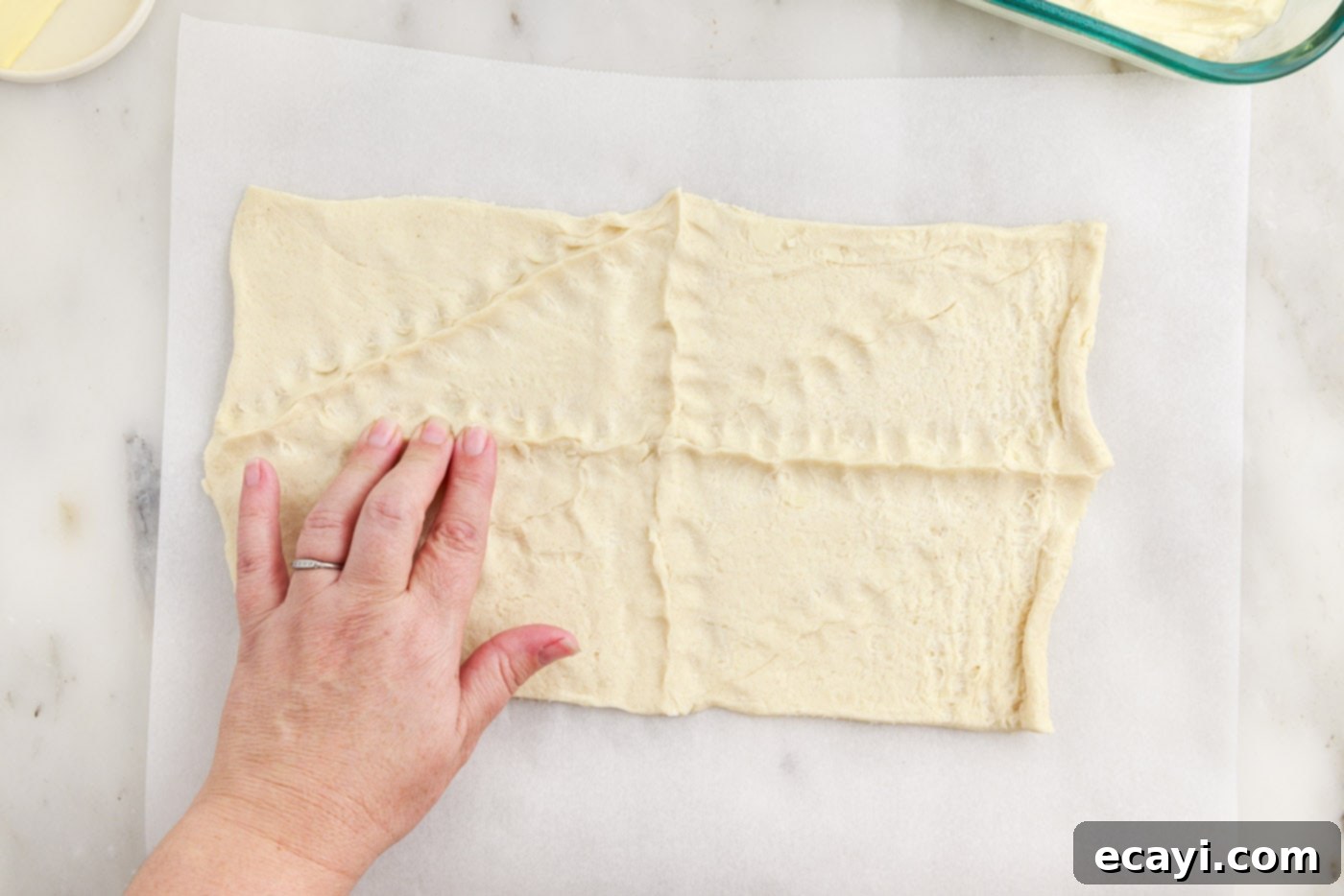 Cinnamon Swirl Sopapilla Cheesecake 10 Hands carefully pressing together the perforations of crescent roll dough on parchment paper to create a unified, smooth sheet for the top crust.