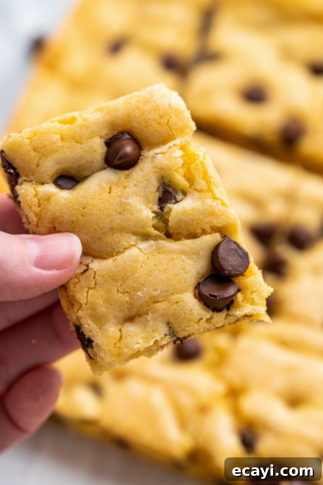 Easy Cake Mix Cookie Bars 13 A hand holding a single, perfectly baked Cake Mix Cookie Bar, showing its soft texture and abundant chocolate chips, with more bars in the background.