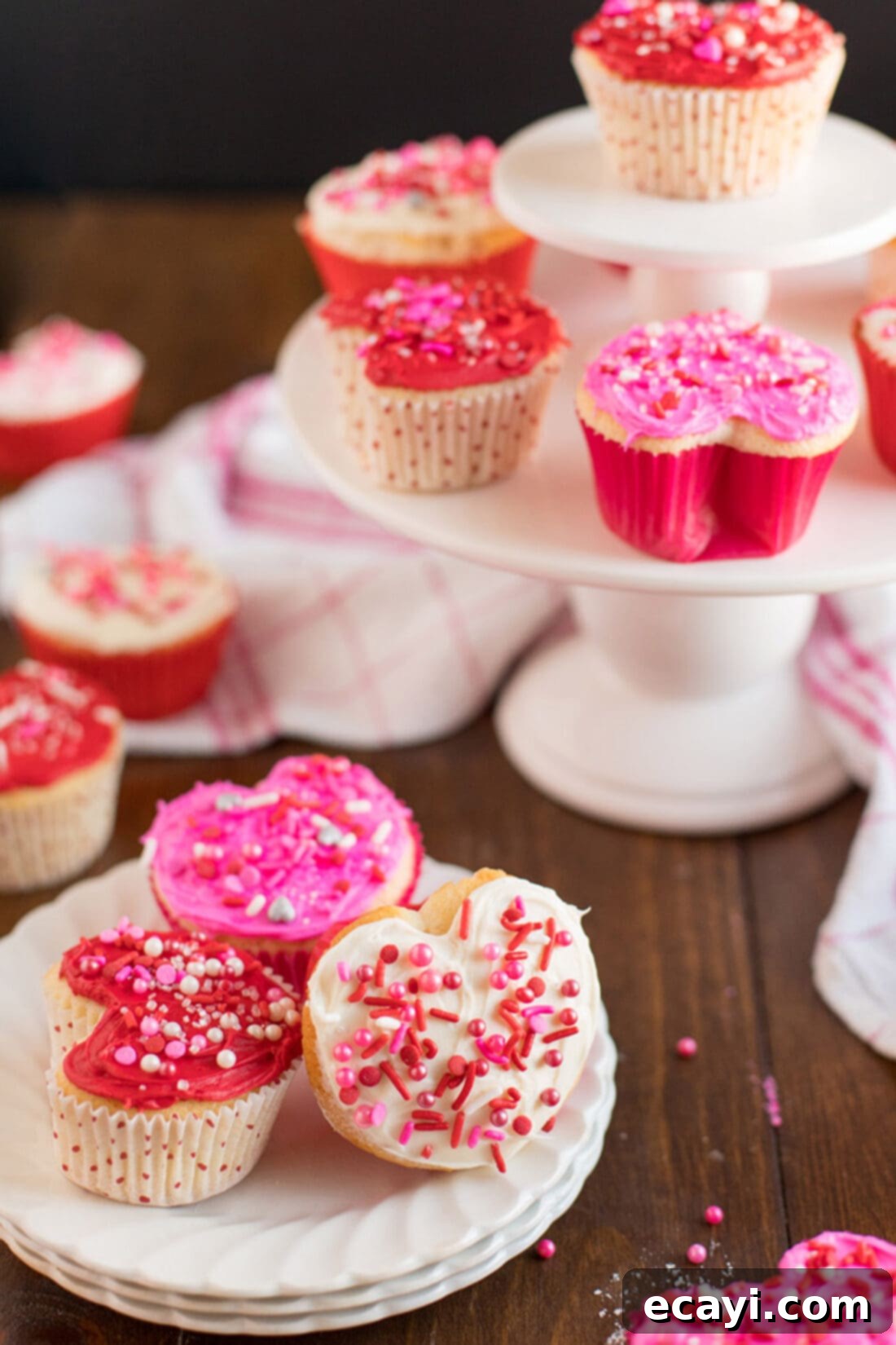 These Valentine cupcakes are shaped like hearts and covered in white, pink and red frosting. Make these super easy heart shaped treats with this recipe. Valentine Heart Cupcakes beautifully arranged on a plate, with additional cupcakes in the background, demonstrating a lovely presentation.