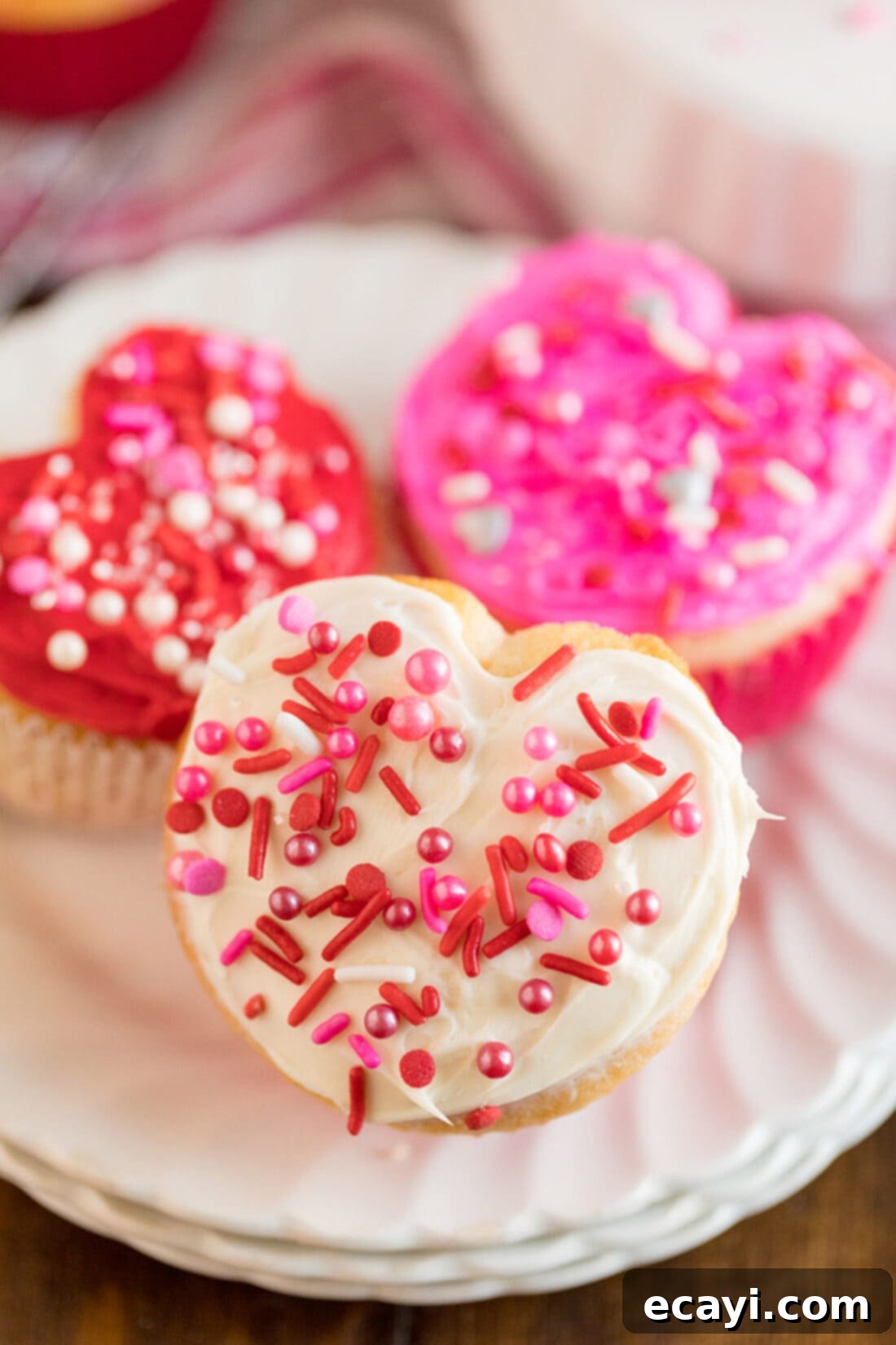 These heart cupcakes are shaped like hearts and covered in white, pink and red frosting. Make these super easy Valentine cupcakes from scratch or a box! #valentinesday #valentinesdaytreats #desserts #love #valentines #cupcakes #cupcakerecipes #cake #anniversary Three beautifully frosted Valentine heart cupcakes displayed on a white plate, showcasing their unique shape and vibrant colors.