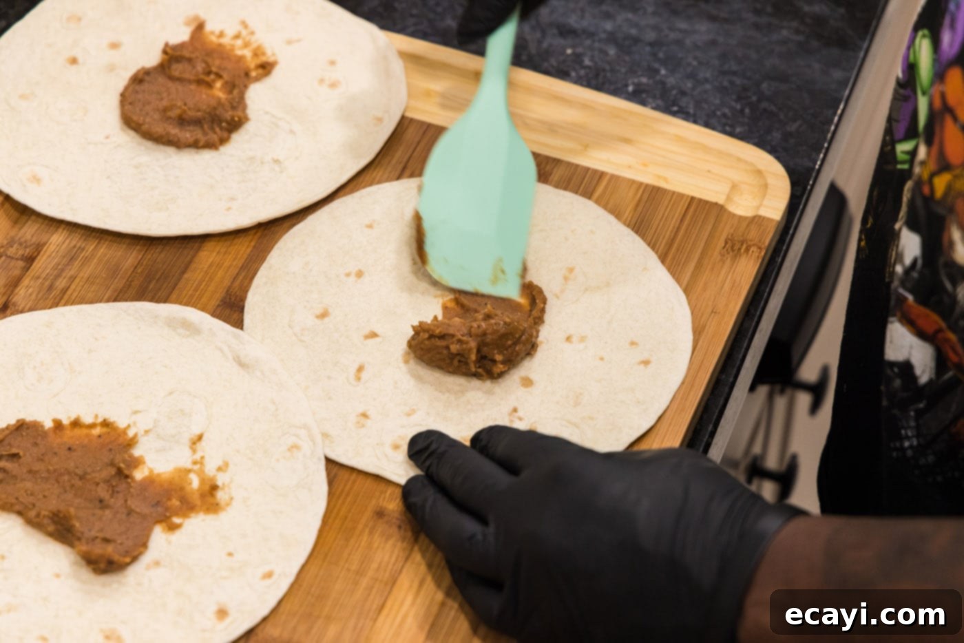 Chimichanga Perfection 11 Spatula spreading refried beans evenly onto the center of a flour tortilla for chimichanga assembly