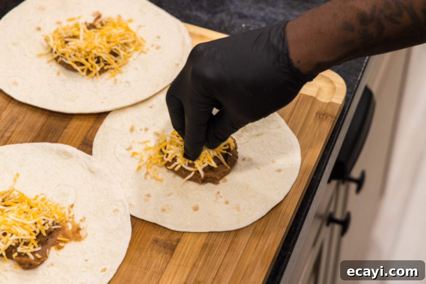 Chimichanga Perfection 12 Shredded cheddar cheese being added on top of refried beans on a tortilla shell