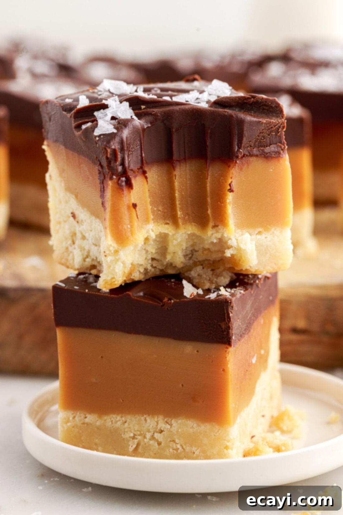 Golden Caramel Indulgence 2 Two Millionaire’s Bars stacked on top of each other with a bite taken out of the top bar, showcasing the three distinct layers: shortbread, caramel, and chocolate ganache.