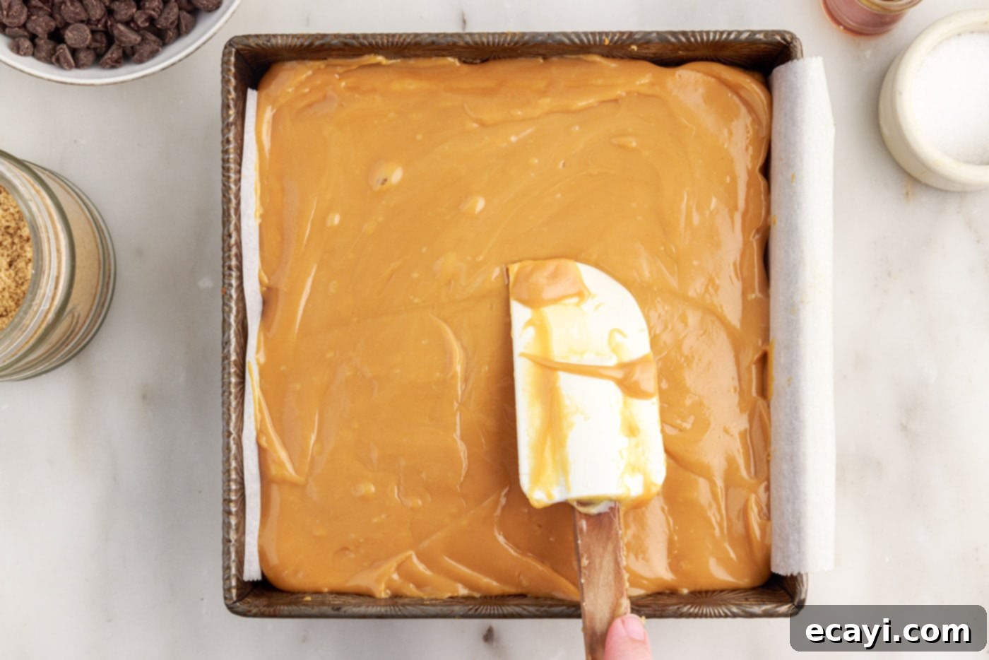 Golden Caramel Indulgence 13 A rubber spatula spreading warm, golden caramel evenly over the baked shortbread crust in a pan.