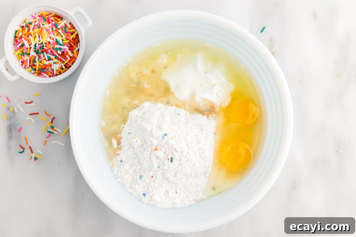 Easy Funfetti Cake Mix Cookies 5 cake mix, eggs, sour cream, and oil being stirred together in a large mixing bowl to form the cookie dough.