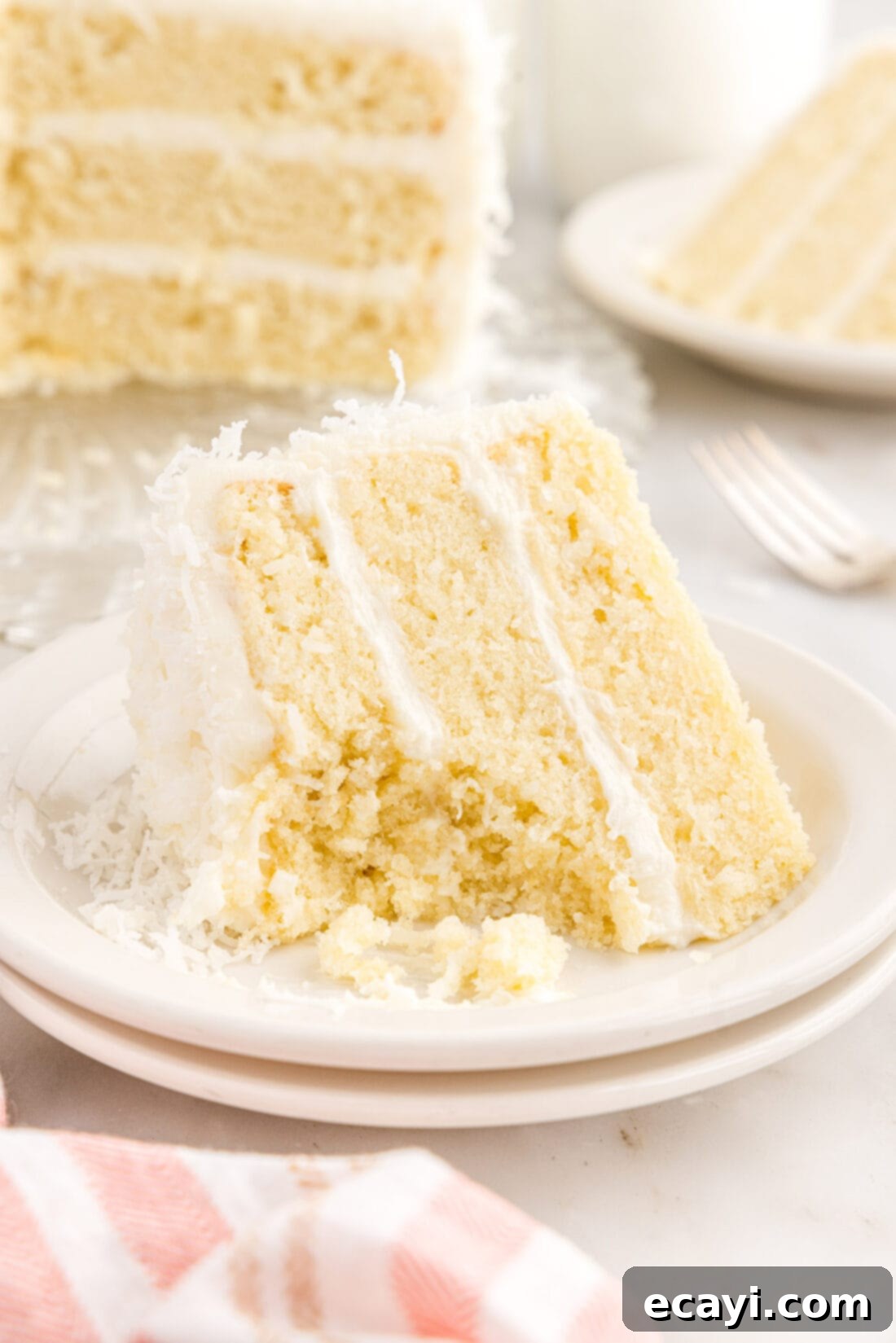 Tropical Cloud Cake 2 Slice of homemade moist coconut cake on a white plate with a bite removed, showcasing fluffy cream cheese frosting and shredded coconut