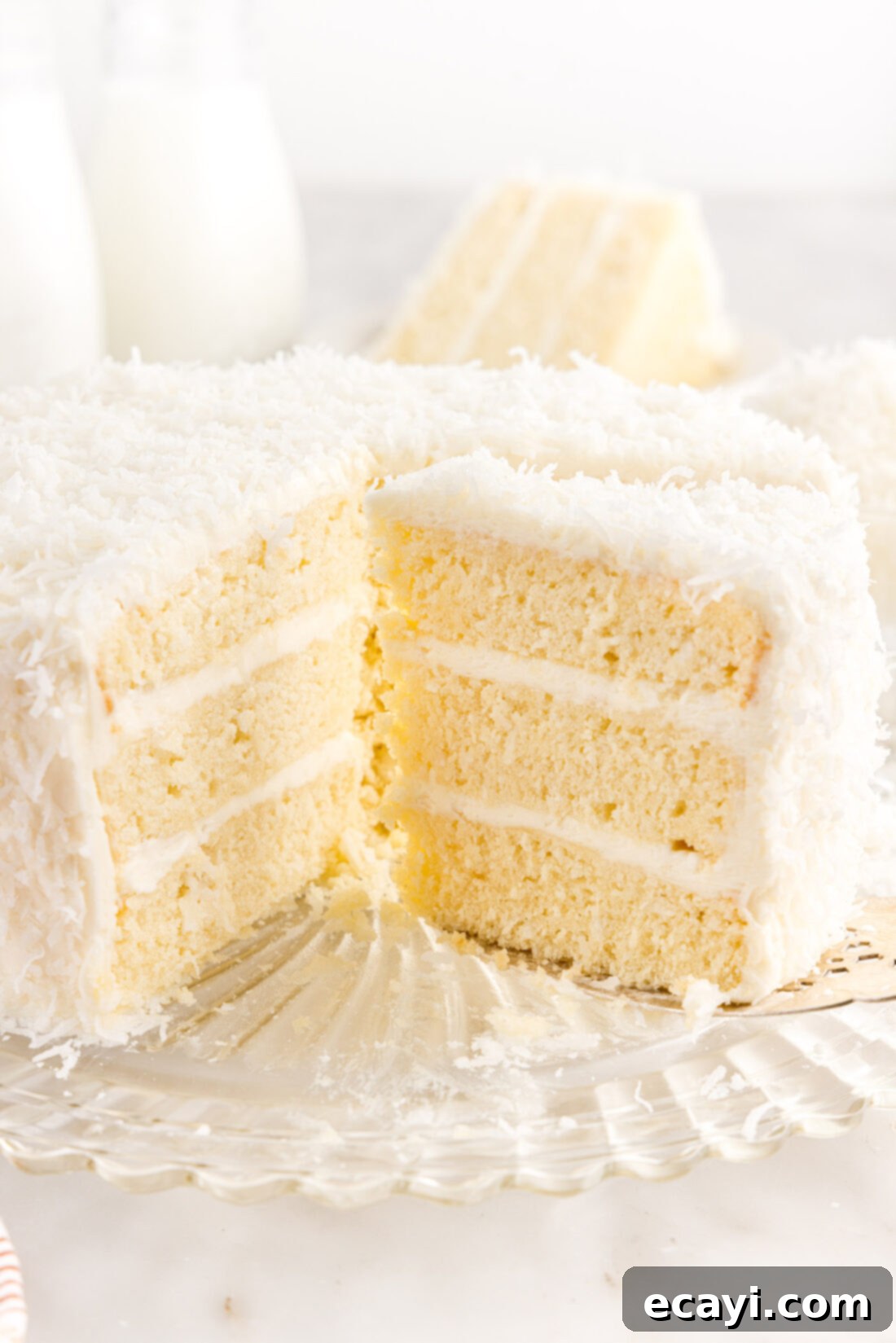 Tropical Cloud Cake 5 Two slices of homemade coconut cake on a white plate, with a full coconut cake in the background, all topped with cream cheese frosting and shredded coconut