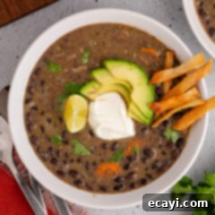 Savory Black Bean Delight 15 A vibrant bowl of black bean soup, generously topped with fresh lime wedges, a dollop of creamy sour cream, slices of ripe avocado, and crunchy tortilla strips, captured from above.