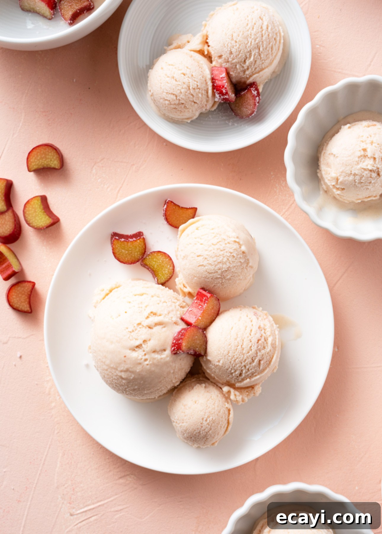 Dairy-Free Vegan Sicilian-Style Gelato Video Recipe 4 Scoops of rhubarb gelato rest on white dishes, elegantly garnished with slices of red and yellow fruit. The backdrop is a peach-colored surface adorned with a few scattered fruit slices. // FoodNouveau.com