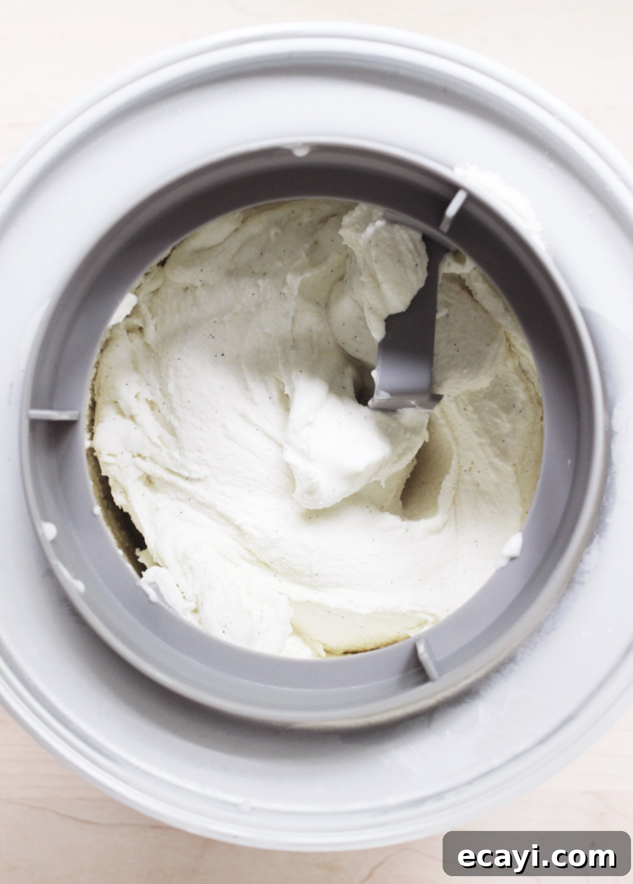 Dairy-Free Vegan Sicilian-Style Gelato Video Recipe 6 Top view of creamy, white Sicilian-Style Gelato being churned in an ice cream maker with a gray paddle. The gelato appears smooth and semi-frozen, showcasing the process of thickening and freezing. // FoodNouveau.com