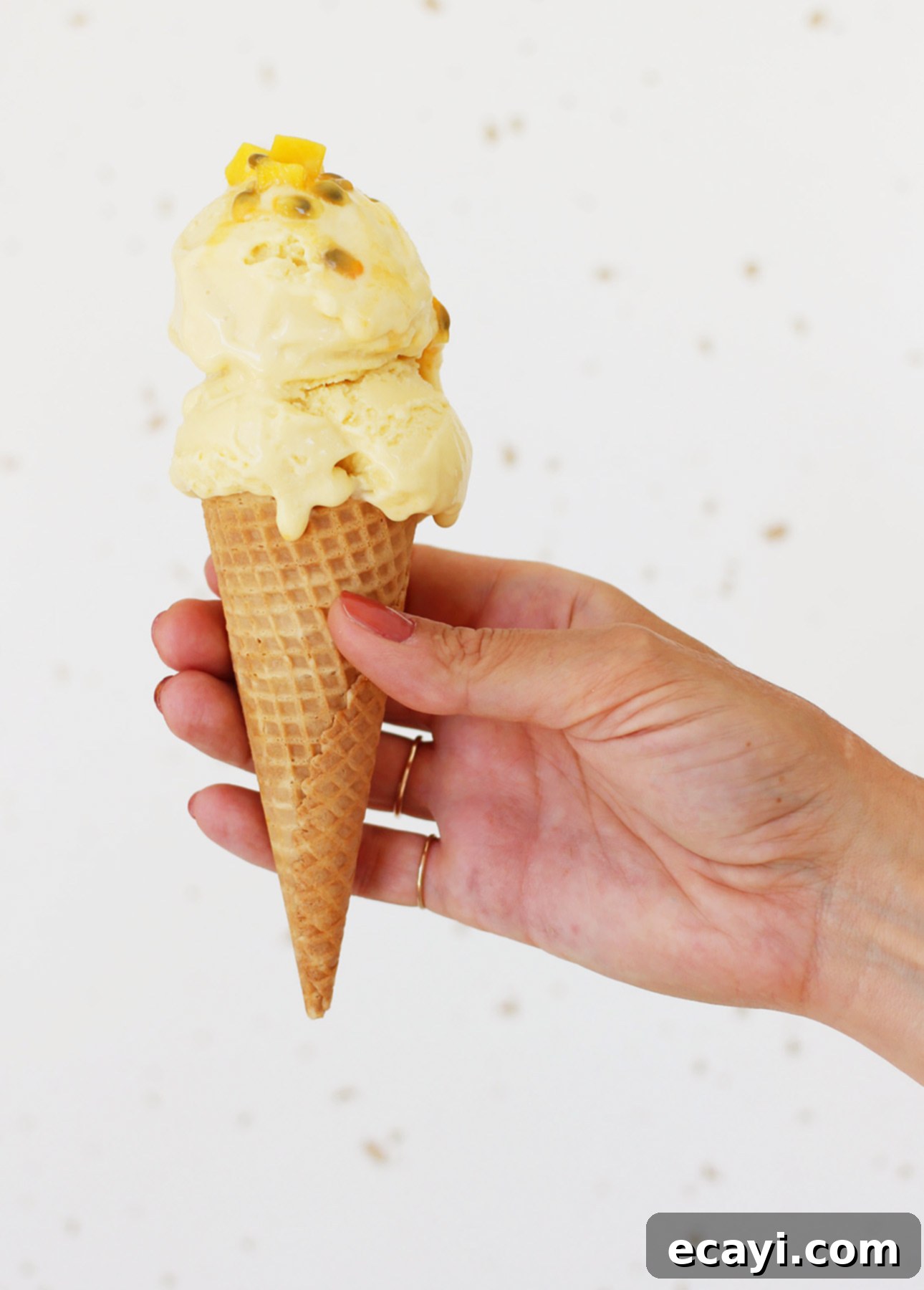 Dairy-Free Vegan Sicilian-Style Gelato Video Recipe 8 A hand holds a cone with two scoops of mango gelato, each topped with small yellow pieces. The creamy texture is complemented by the persons painted fingernails. In the background lies a light, speckled surface. // FoodNouveau.com