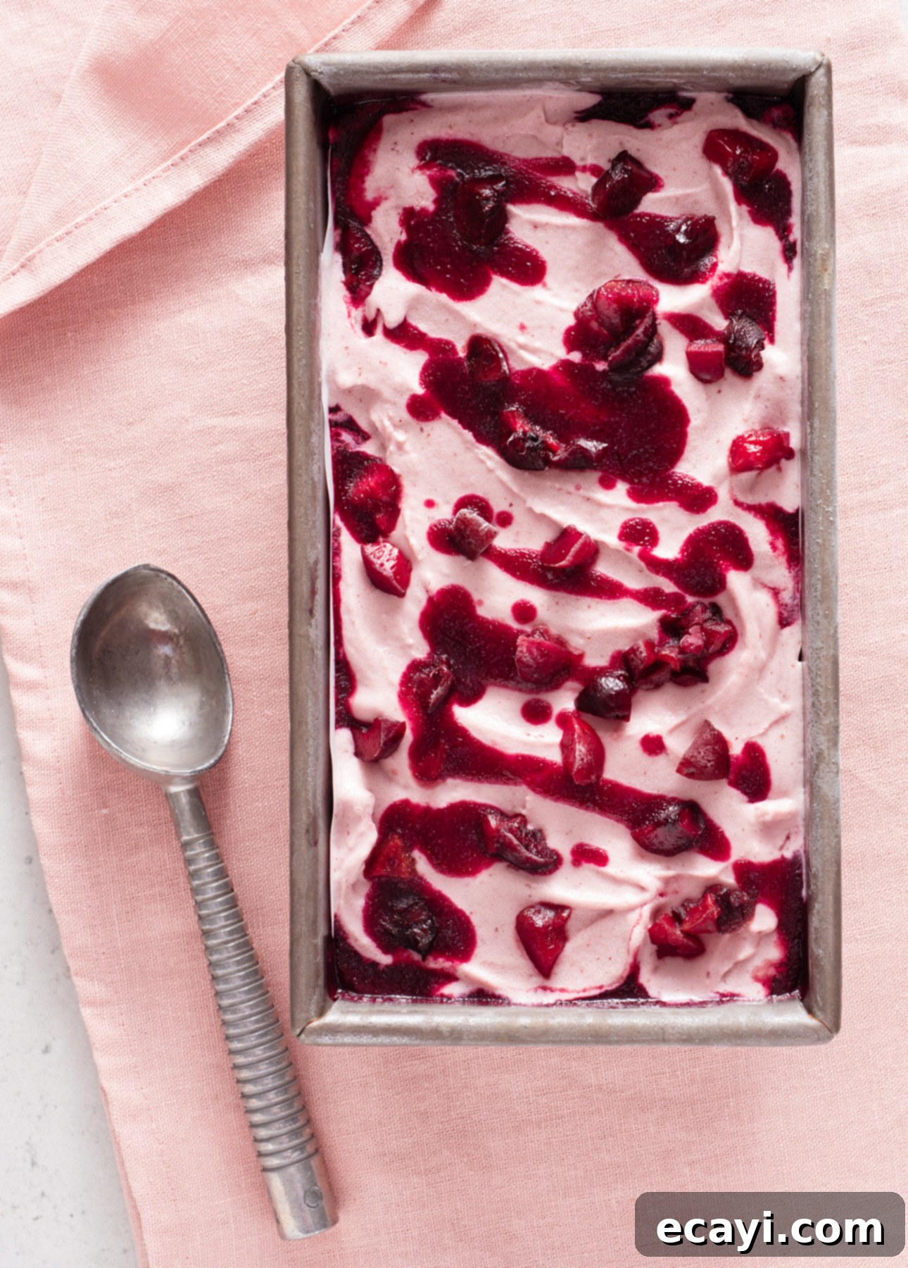 Luscious Cherry Gelato 3 A gray loaf pan filled with beautifully swirled cherry gelato, showcasing creamy pink tones marbled with rich dark red cherry puree. A vintage silver spoon lies gracefully beside the pan on a soft pink cloth, highlighting the tempting texture and visual appeal of the homemade dessert.
