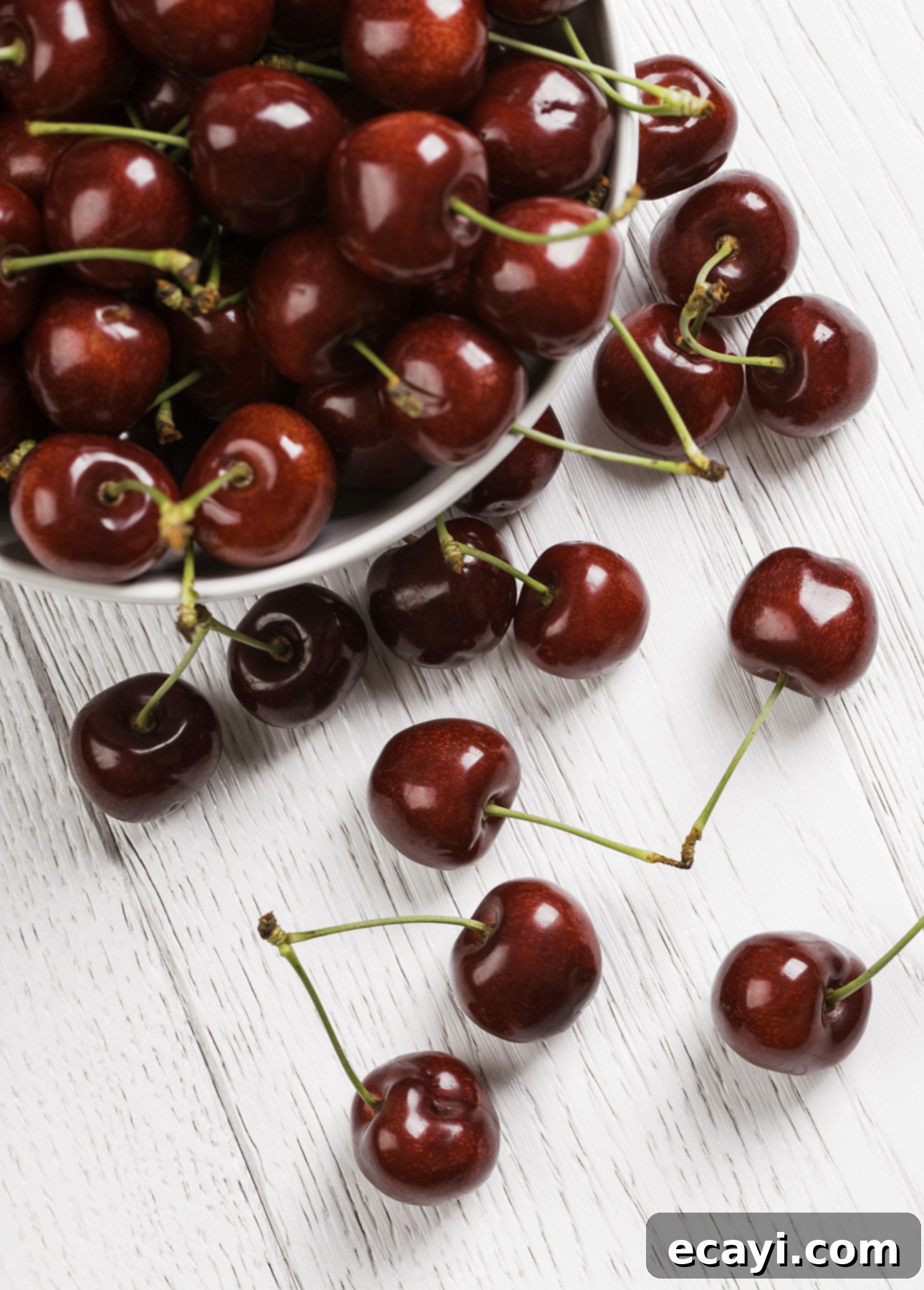 Luscious Cherry Gelato 4 A pristine white bowl filled with glossy, dark red cherries, vibrant and fresh, is gently tipped over on a clean white wooden surface. Several cherries have spilled out, scattering onto the table, with a few showcasing their vivid green stems, emphasizing their freshness and natural beauty.