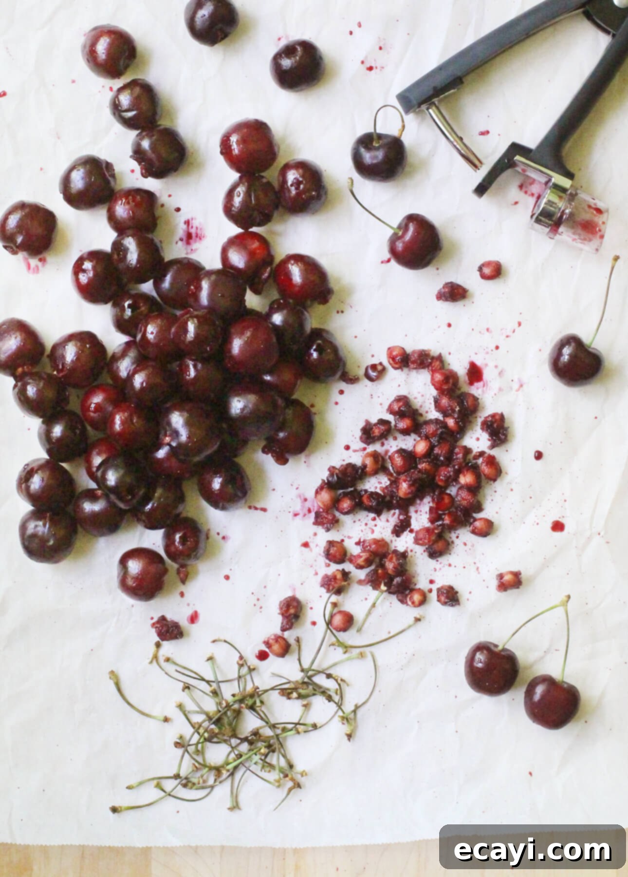 Luscious Cherry Gelato 5 Fresh, vibrant cherries are artfully scattered across a clean white surface, some whole and unblemished, others perfectly pitted. A sleek cherry pitter, a true kitchen hero, is positioned on the right, accompanied by small, neat piles of removed pits and delicate stems, showcasing the efficiency of the tool and the readiness of the fruit for culinary creation.