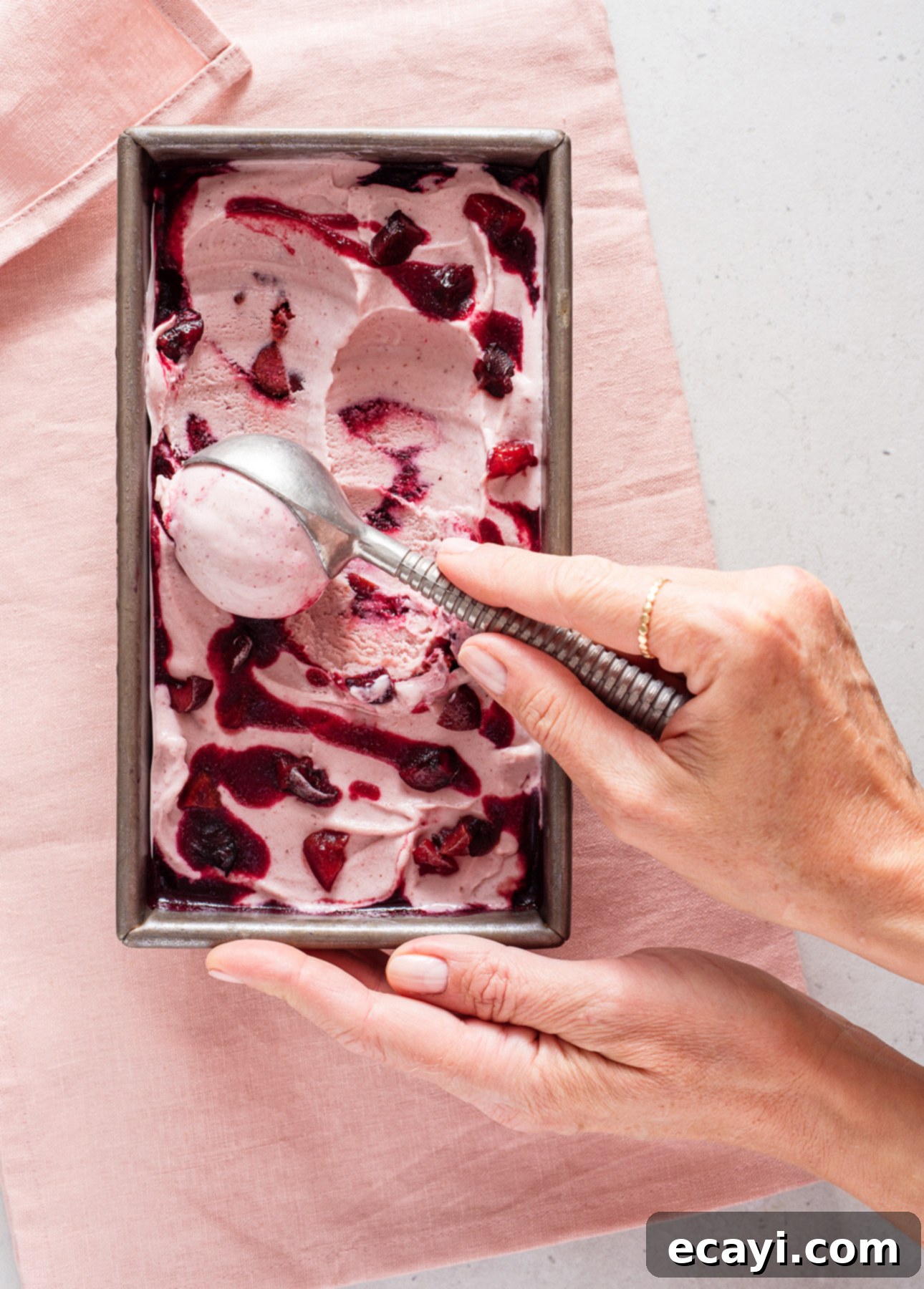 Luscious Cherry Gelato 6 A hand gently holds a metal ice cream scooper, expertly serving vibrant pink cherry gelato from a rectangular metal container, which is elegantly placed on a soft pink cloth. The gelato glistens with beautiful, dark red swirls of cherry puree, indicating its rich fruit flavor and appealing texture.