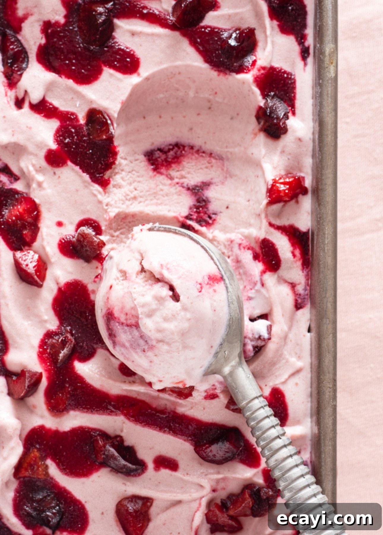 Luscious Cherry Gelato 7 A gleaming metal ice cream scooper rests invitingly in a tray of smooth, pink Cherry Gelato. The surface is beautifully adorned with vivid swirls of dark red cherry puree and scattered pieces of fresh fruit, creating a striking visual contrast. The rich, creamy texture of the gelato promises a delightful indulgence.