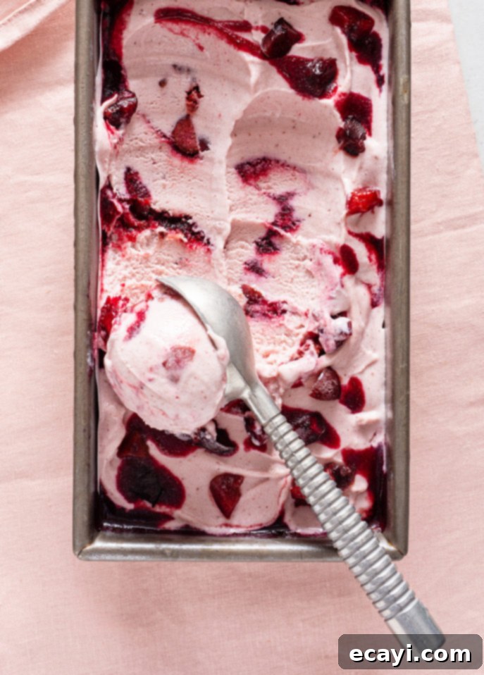 Luscious Cherry Gelato 8 A rectangular metal container brims with Cherry Ripple Gelato, creamy pink and swirled with dark red berry preserves and chunks. A silver spoon rests on top, all set against a soft pink fabric backdrop. // FoodNouveau.com