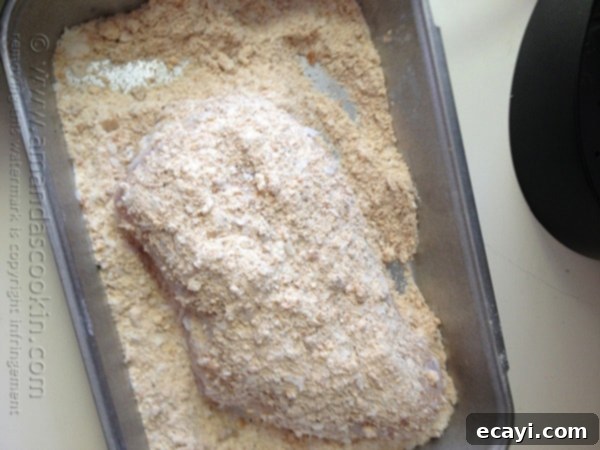 Golden-Crisp Oven Chicken Breasts 9 An overhead photo of a raw chicken breast being coated thoroughly in the crumb mixture in a shallow dish.