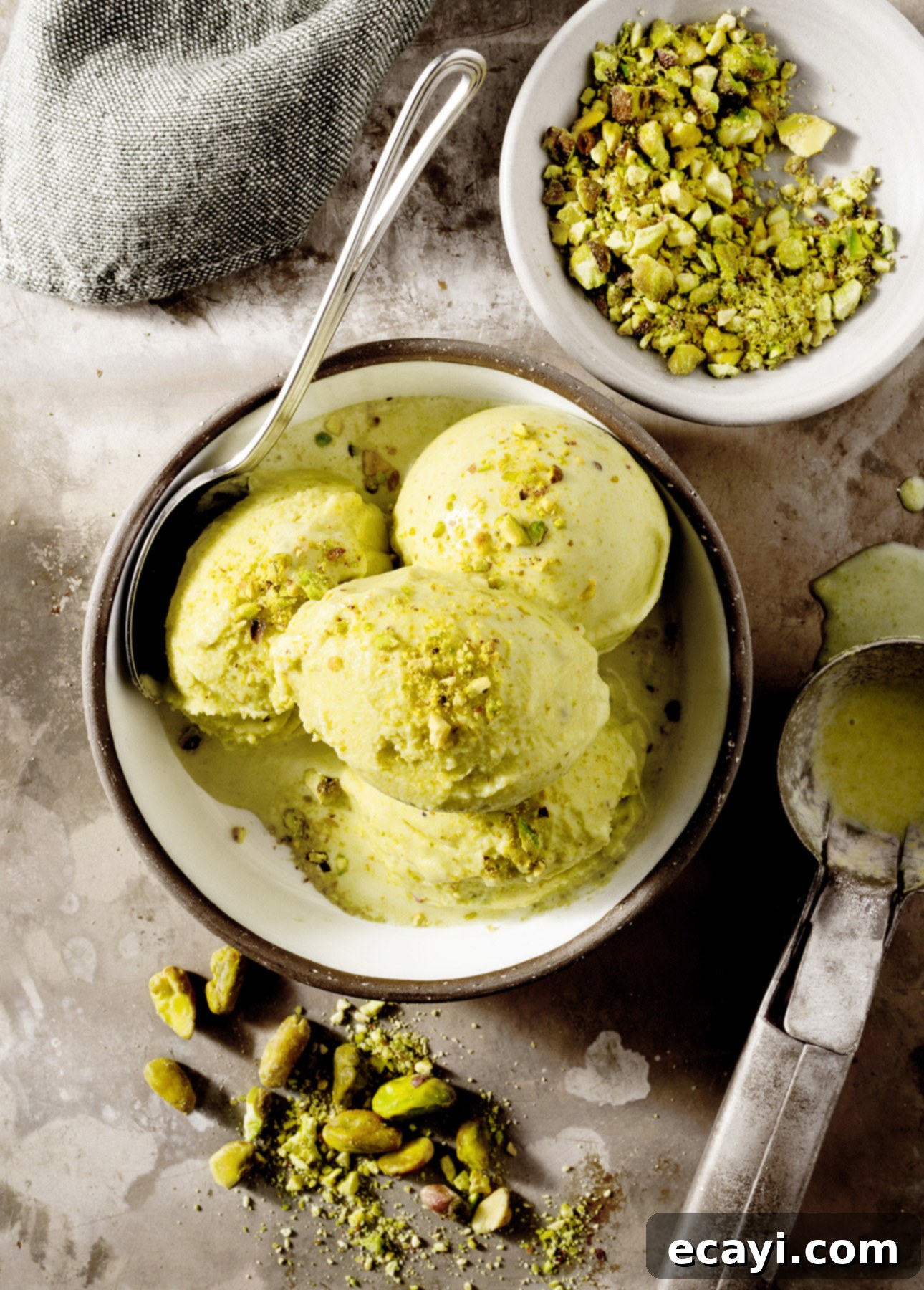 Authentic Pistachio Gelato from Our Homemade Paste 10 A delightful bowl of pistachio gelato featuring three perfectly sculpted scoops, elegantly garnished with a sprinkle of crushed pistachios. A sleek metal spoon rests invitingly in the bowl beside a small dish of chopped pistachios and a vintage ice cream scoop on a textured surface, ready for enjoyment. // FoodNouveau.com