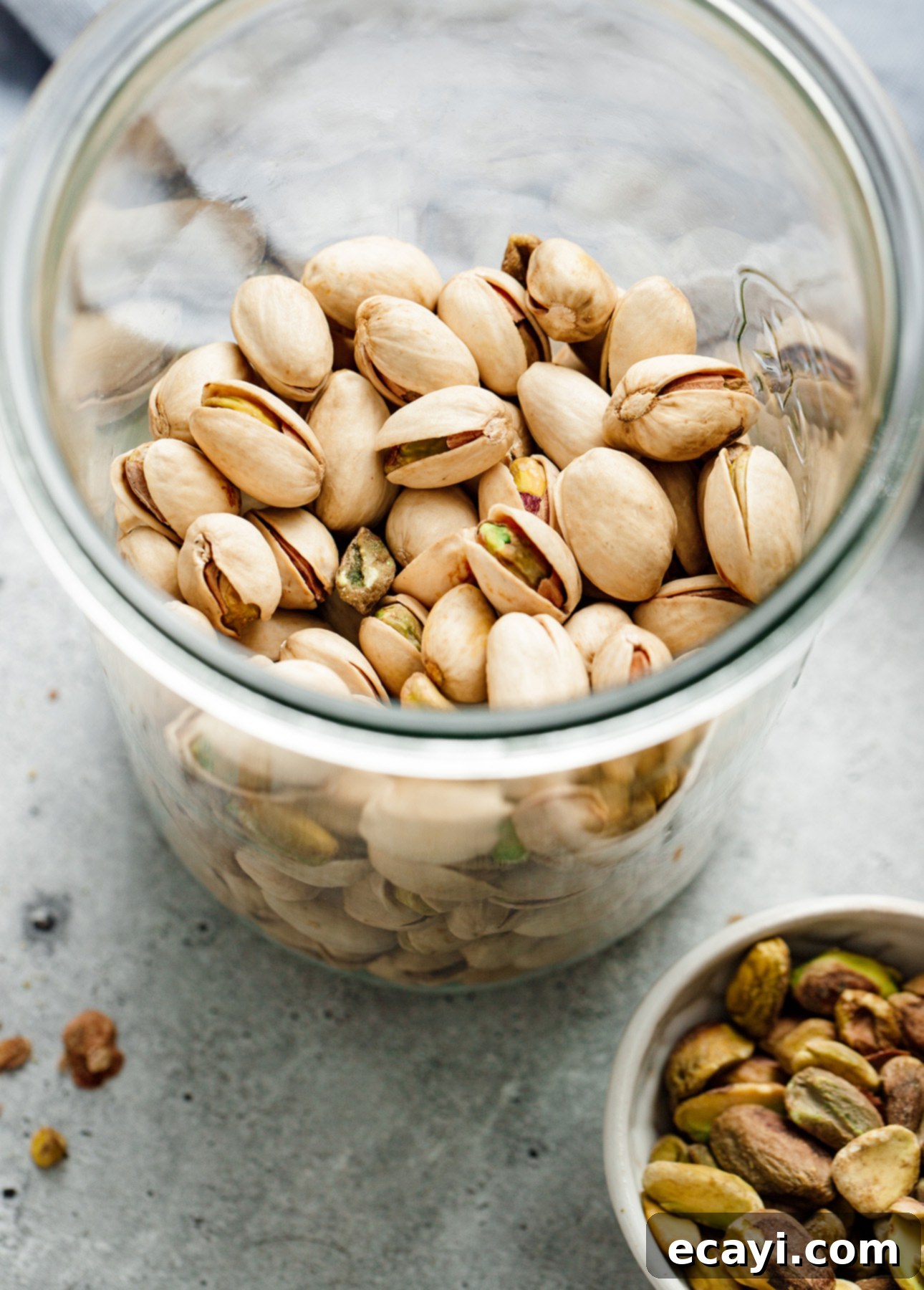 Authentic Pistachio Gelato from Our Homemade Paste 4 A beautifully arranged jar of shelled pistachios on a gray surface, accompanied by a small white bowl holding both shelled and unshelled nuts, with a few pieces scattered nearby. This image highlights the quality ingredients used for homemade pistachio gelato. // FoodNouveau.com