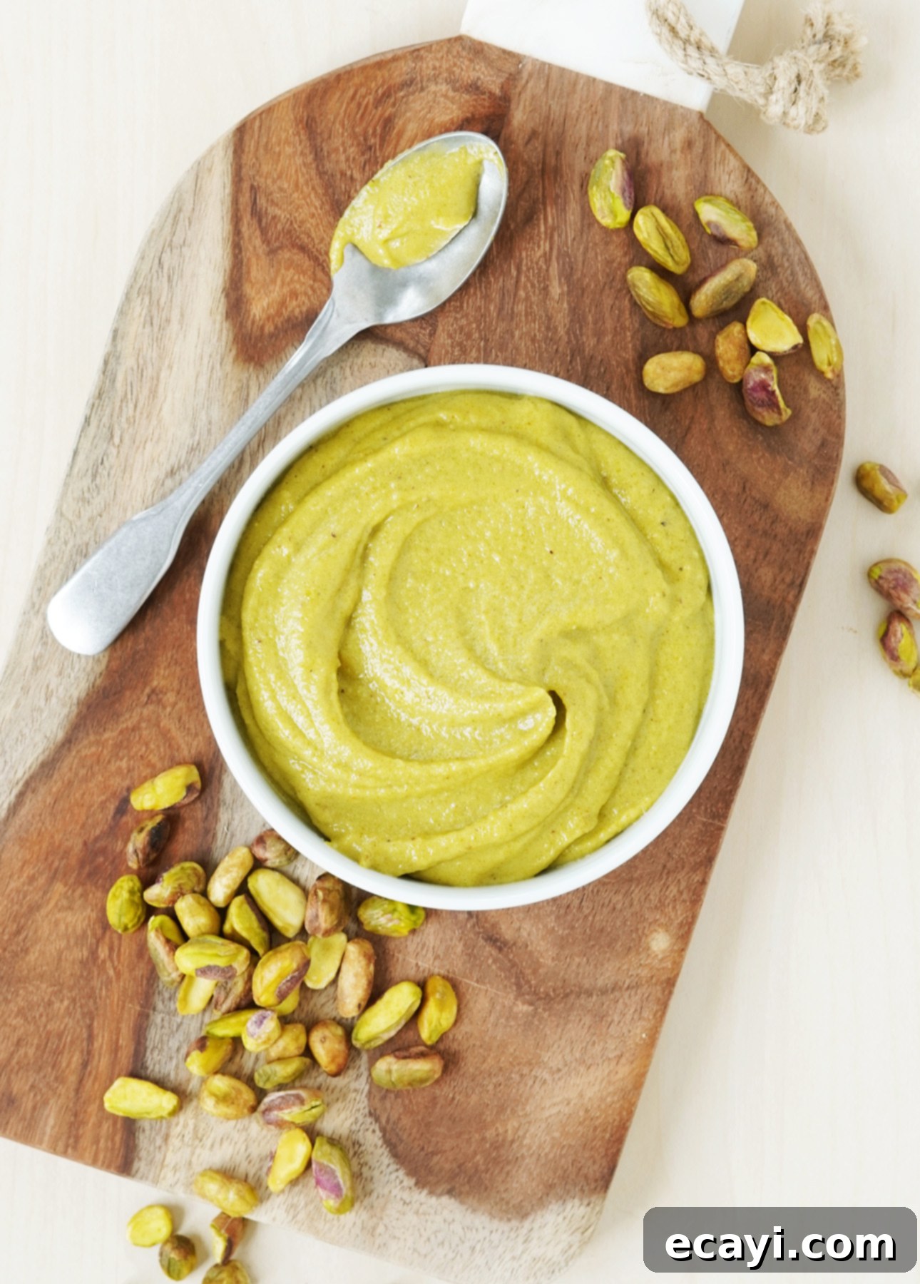 Authentic Pistachio Gelato from Our Homemade Paste 5 A bowl of creamy, bright green homemade pistachio paste rests on a rustic wooden cutting board. A spoon, still coated with some of the rich paste, lies nearby, surrounded by scattered shelled pistachios, emphasizing the key ingredient for this gelato recipe. // FoodNouveau.com