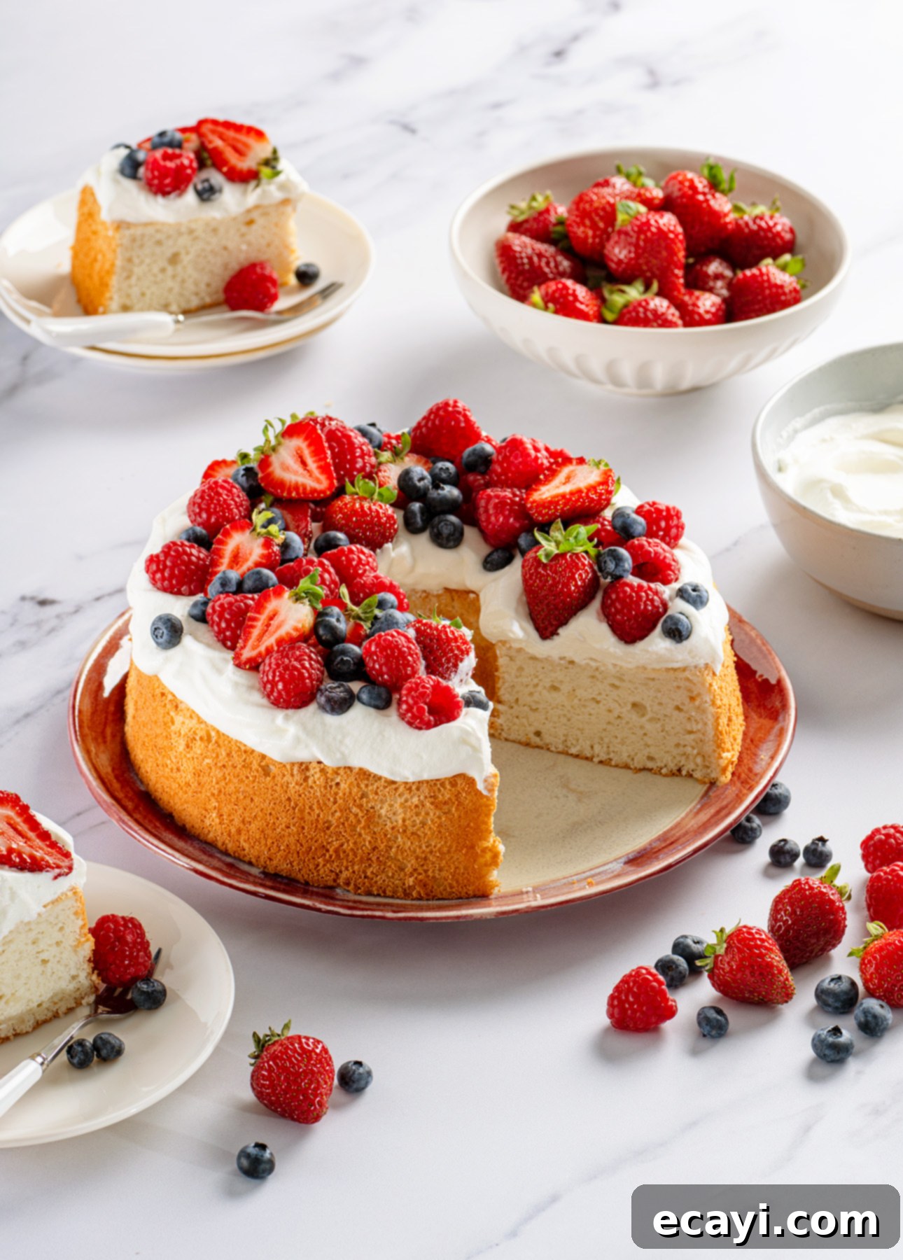 Heavenly Homemade Angel Food Cake 2 A homemade Angel Food Cake topped with whipped cream, strawberries, blueberries, and raspberries sits on a plate. Slices are served on smaller plates nearby, with bowls of fresh berries and whipped cream in the background. // FoodNouveau.com