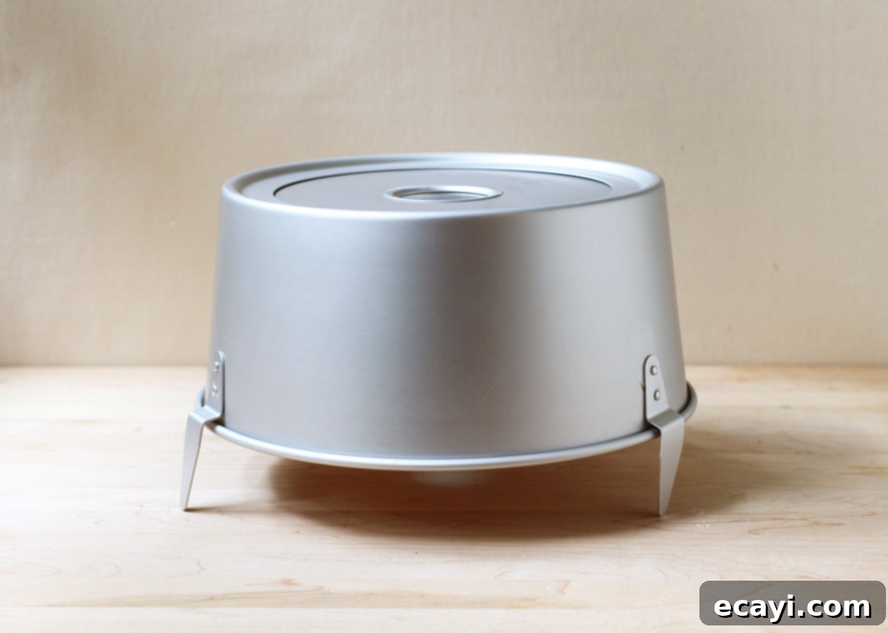 Heavenly Homemade Angel Food Cake 15 A round, silver metal angel food cake pan stands upside down on three foldable legs, placed on a light wooden surface against a plain beige background. // FoodNouveau.com
