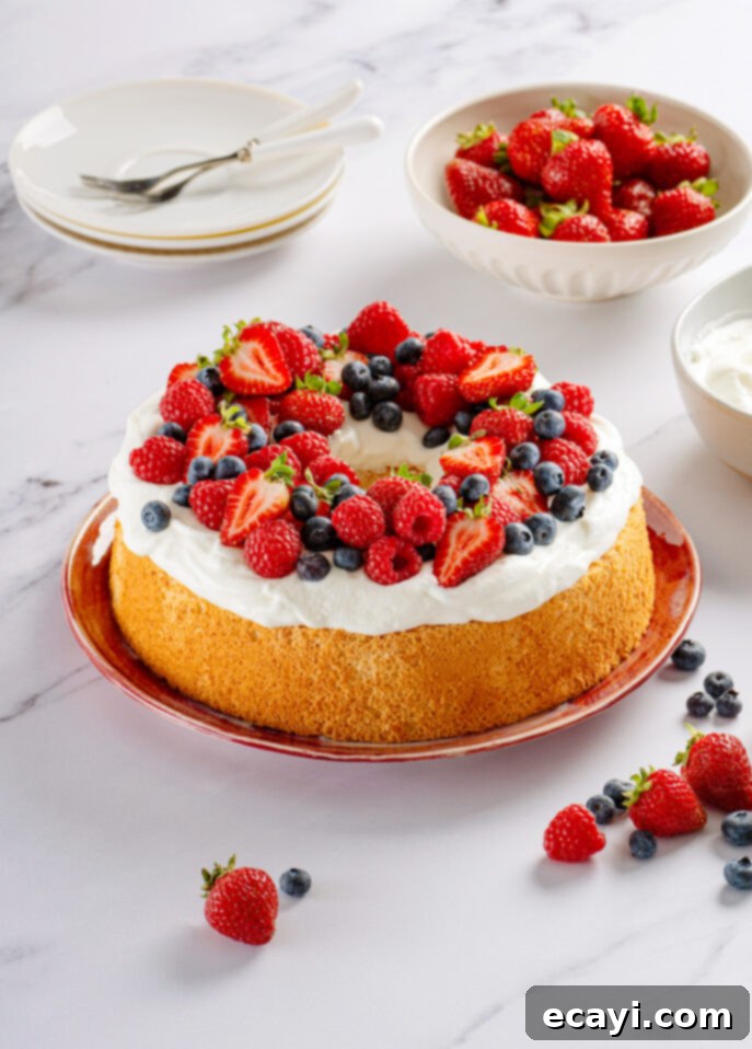 Heavenly Homemade Angel Food Cake 16 An Angel Food Cake topped with whipped cream, strawberries, raspberries, and blueberries sits on a red plate. Nearby are bowls of fresh strawberries, whipped cream, and stacked plates with forks. // FoodNouveau.com