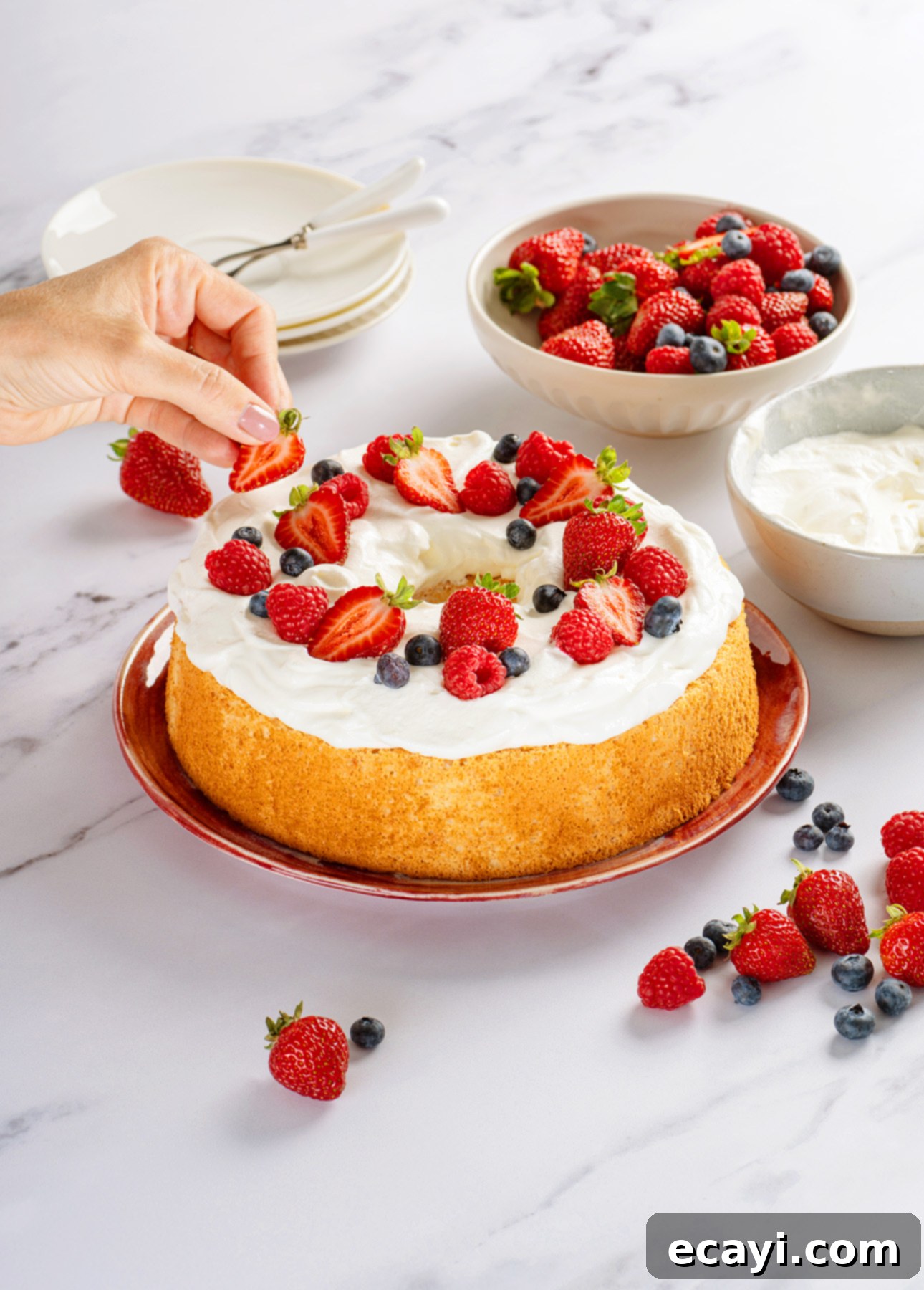 Heavenly Homemade Angel Food Cake 3 A hand places a strawberry on an Angel Food Cake topped with whipped cream and assorted berries. Nearby are bowls of berries, a bowl of whipped cream, and stacked plates with forks on a marble surface. // FoodNouveau.com