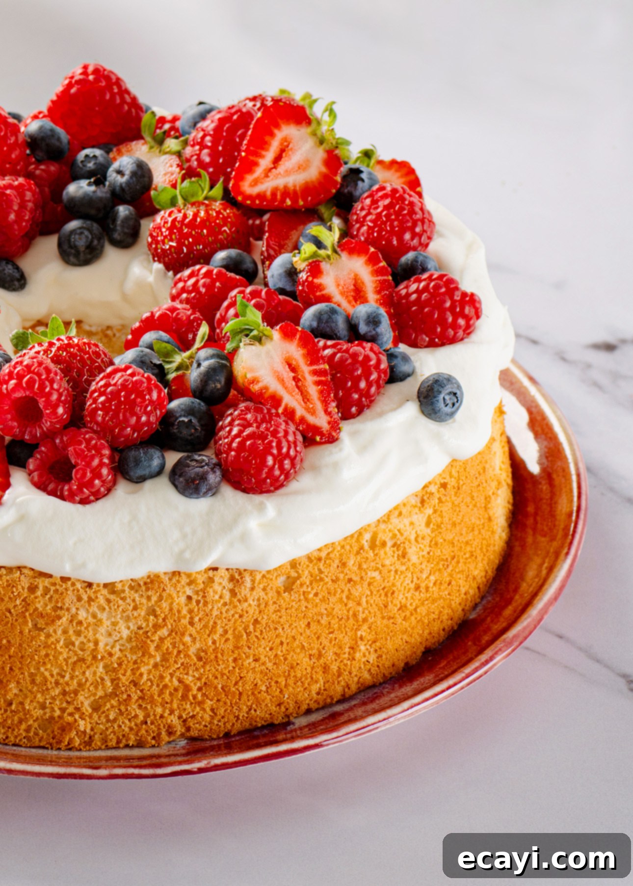 Heavenly Homemade Angel Food Cake 4 A fluffy Angel Food Cake topped with whipped cream and decorated with fresh strawberries, raspberries, and blueberries, served on a red plate against a marble background. // FoodNouveau.com