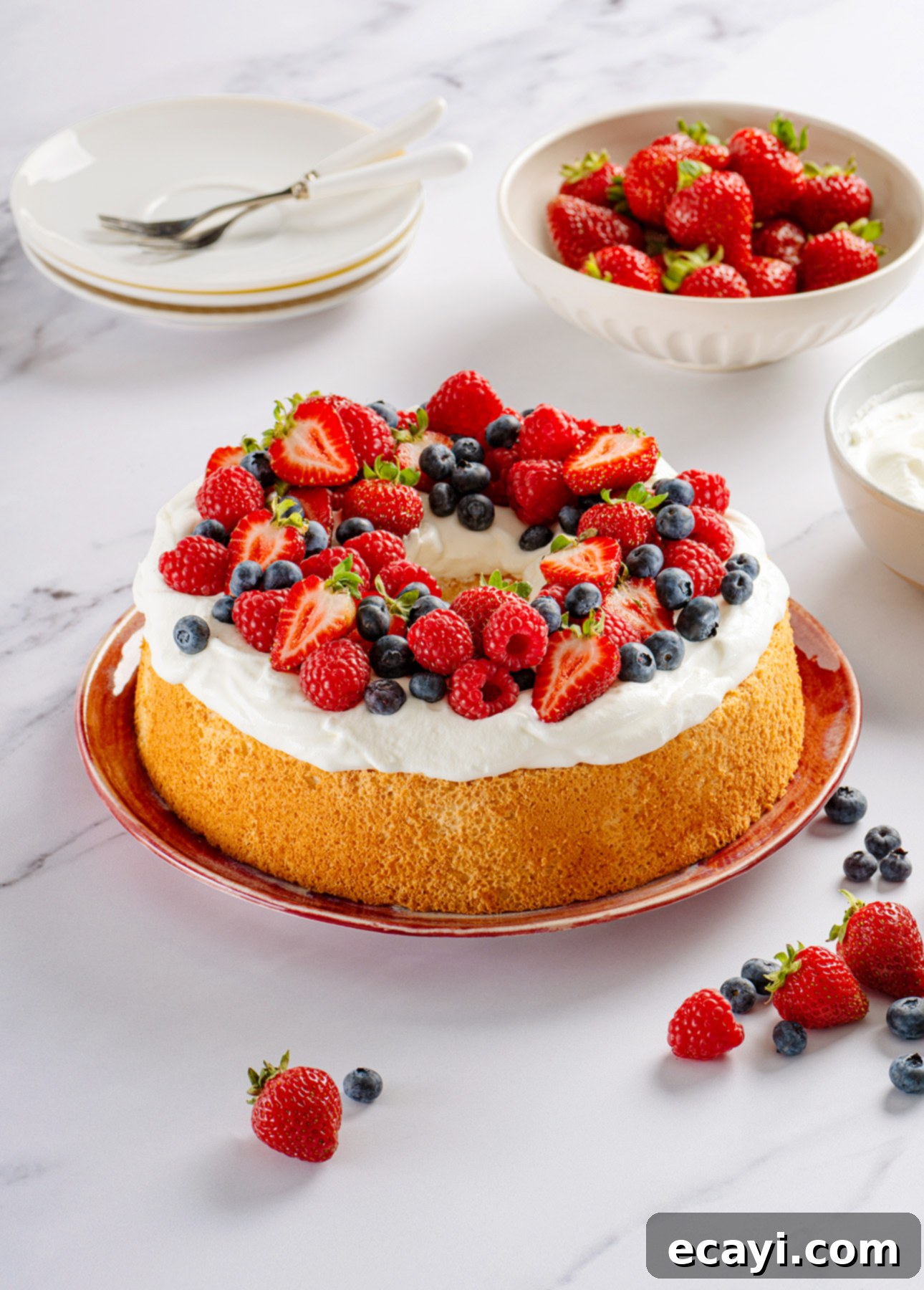 Heavenly Homemade Angel Food Cake 7 An Angel Food Cake topped with whipped cream, strawberries, raspberries, and blueberries sits on a red plate. Nearby are bowls of fresh strawberries, whipped cream, and stacked plates with forks. // FoodNouveau.com