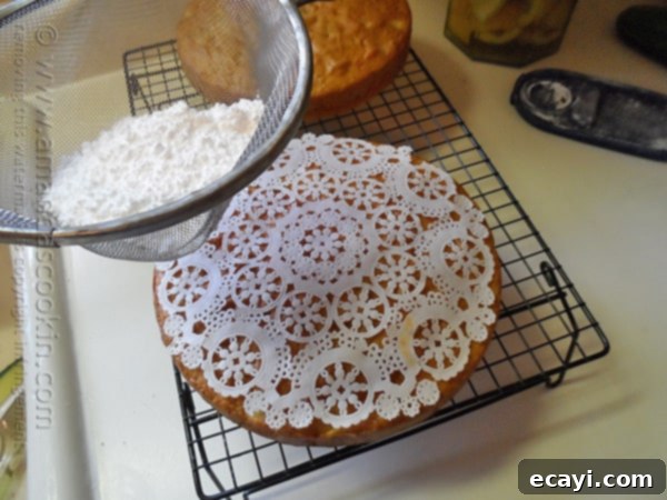 Classic Merryfield Apple Cake 12 A person carefully sprinkling powdered sugar over a Merryfield apple cake using a paper doily as a stencil, creating an intricate design on the cake's surface.