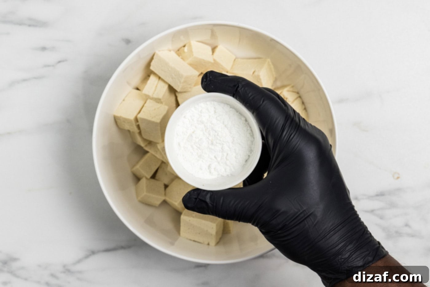 hand holding a bowl of cornstarch over cubed tofu in a bowl