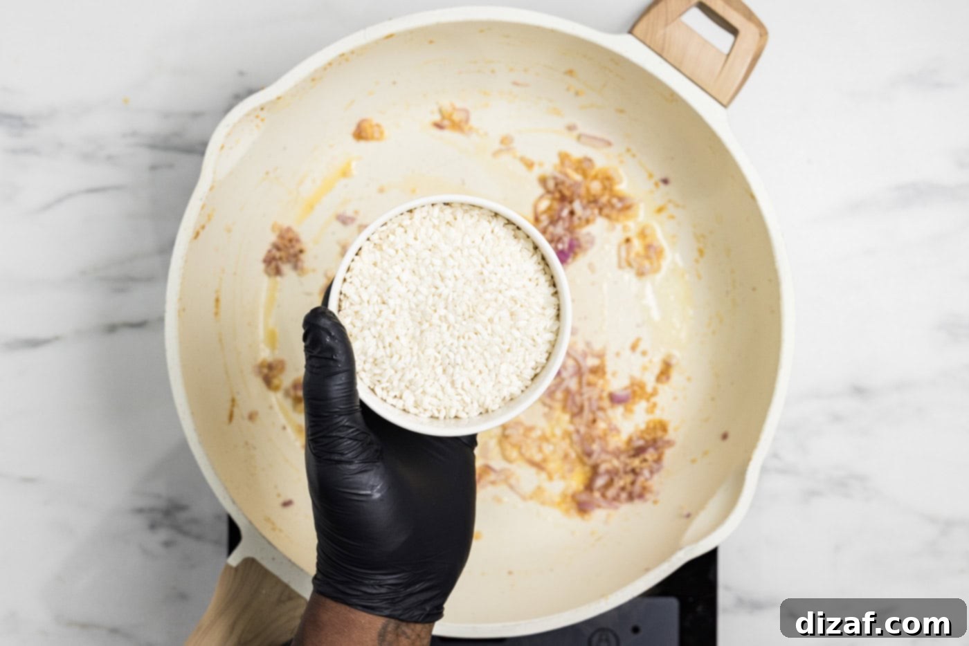 hand holding a bowl of risotto over a skillet with onions and garlic
