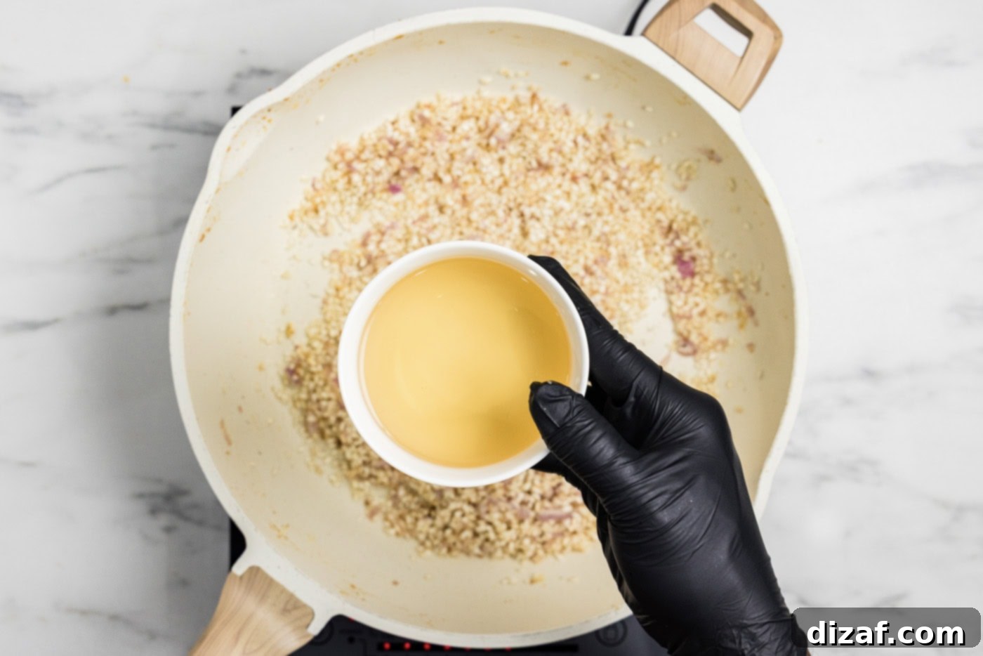 hand holding a bowl of white wine over a skillet of risotto, garlic, and onion